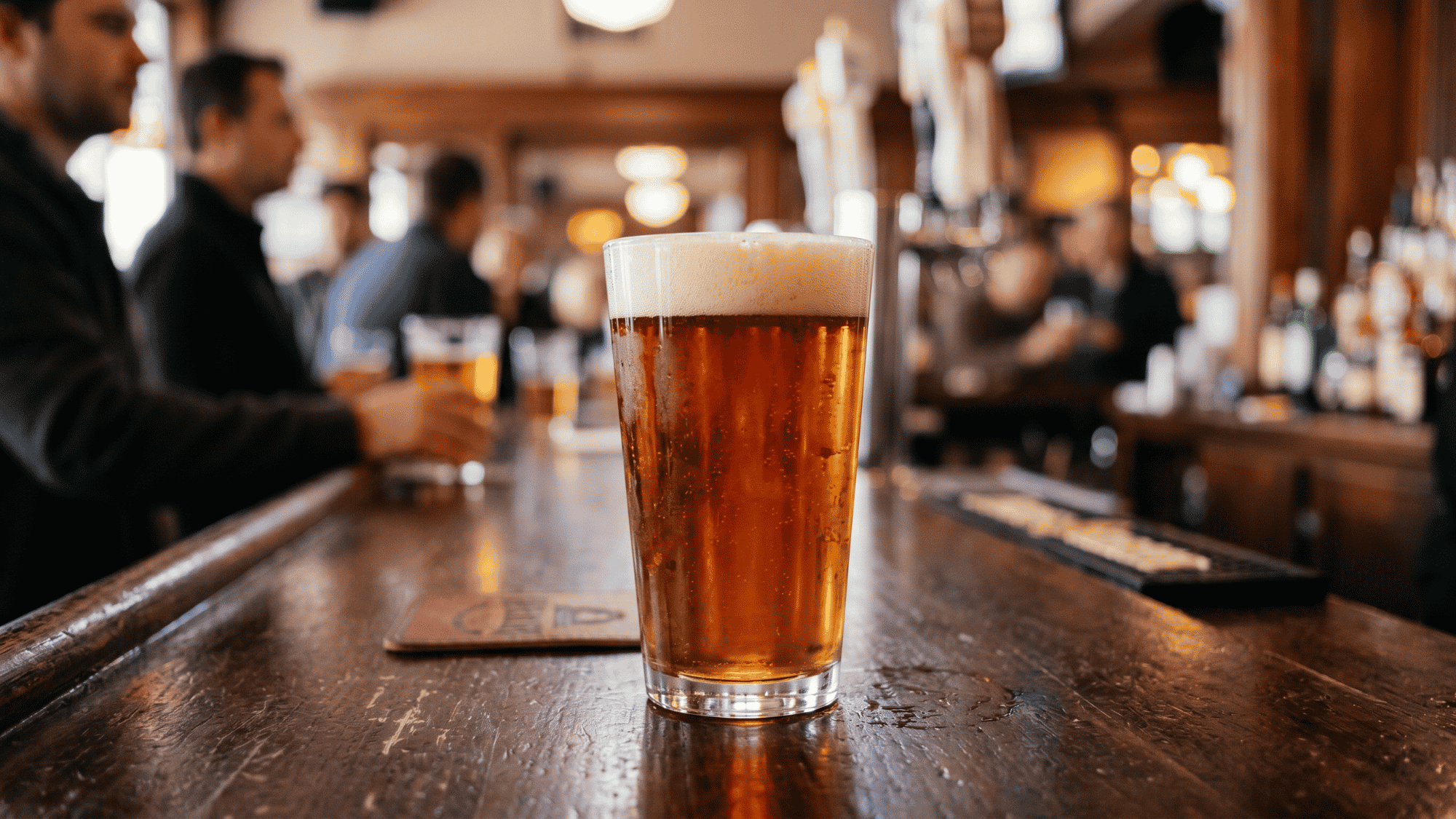 Close up of an American pint glass filled with amber beer in a busy pub bar pub.