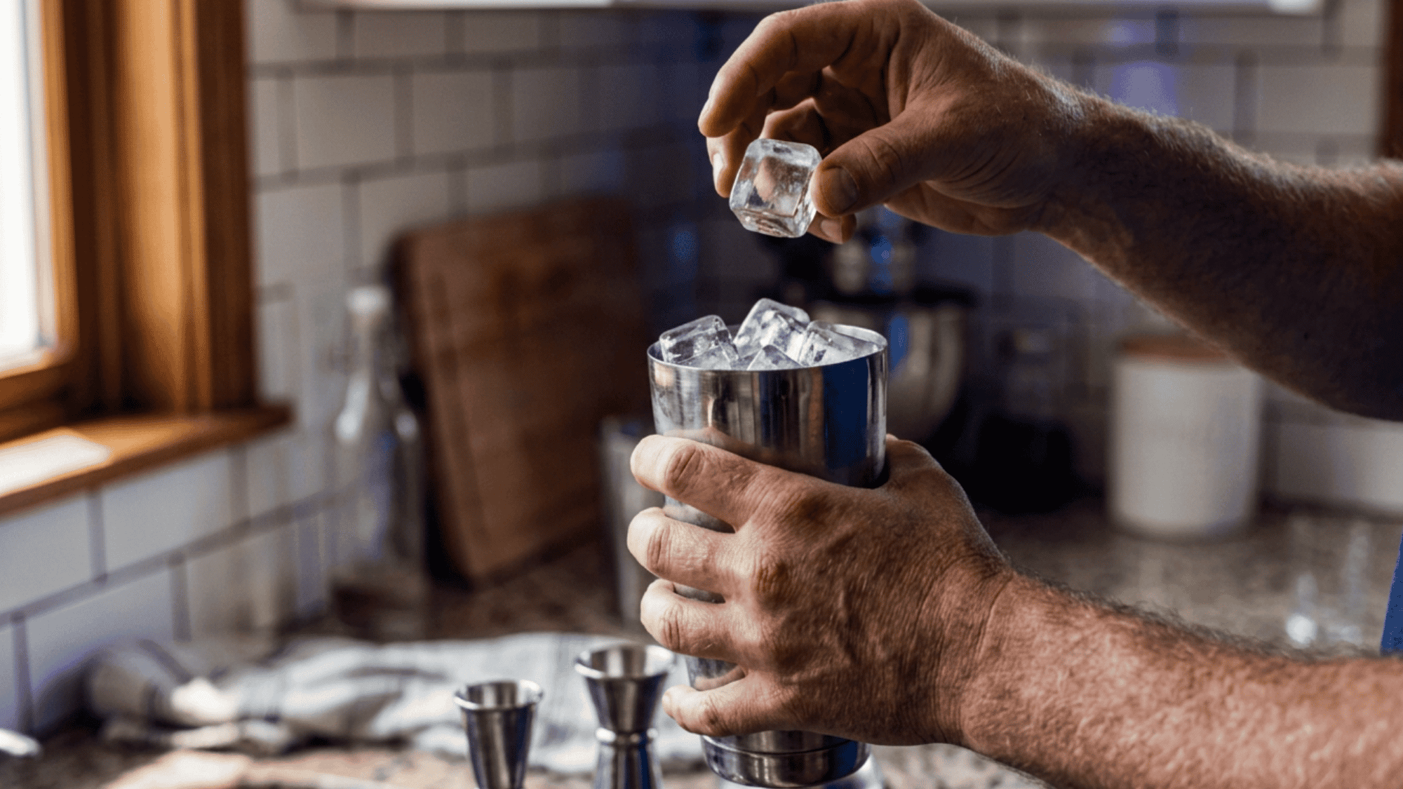 Adding ice cubes into an open stainless steel cocktail shaker to chill the drink.
