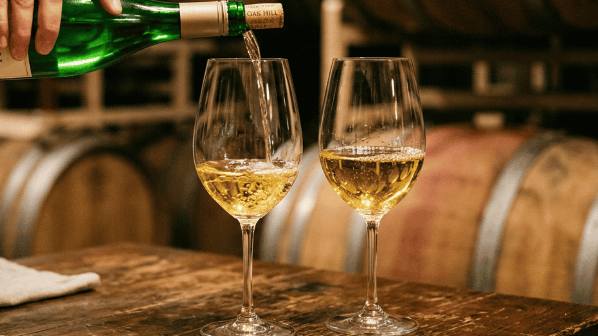 White wine poured into two glasses in a winery with oak barrels in the background