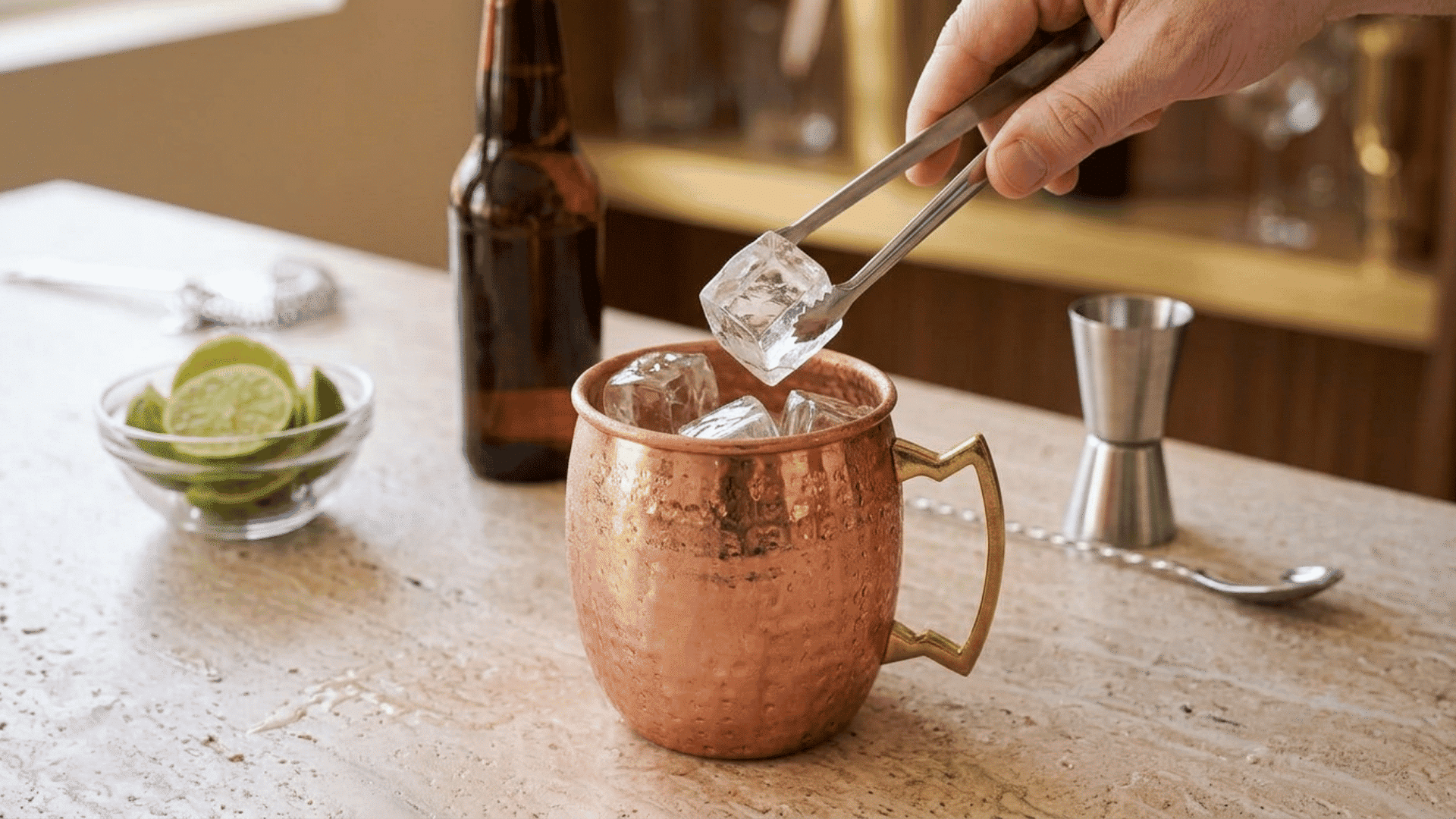 Hand placing a clear ice cube into a copper mug using metal tongs.