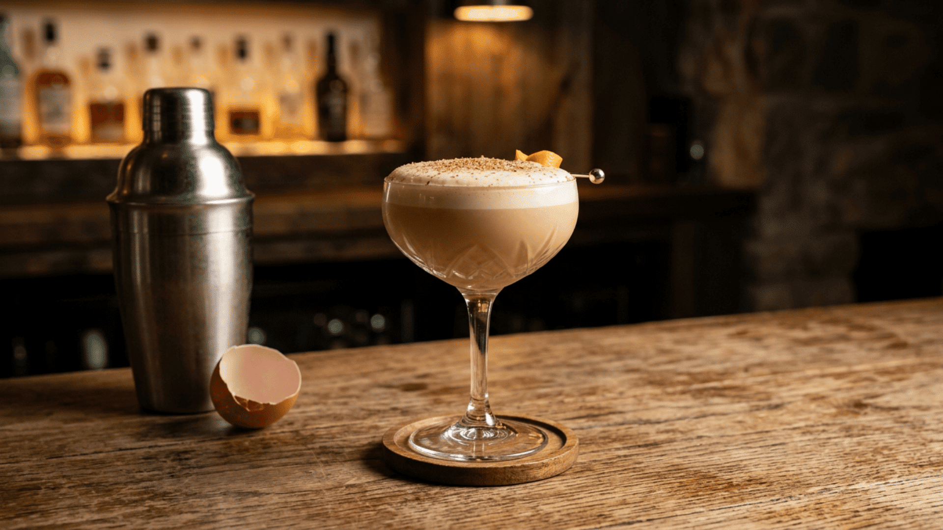 Flip cocktail with foam, nutmeg, shaker, and eggshell on a wooden bar counter.