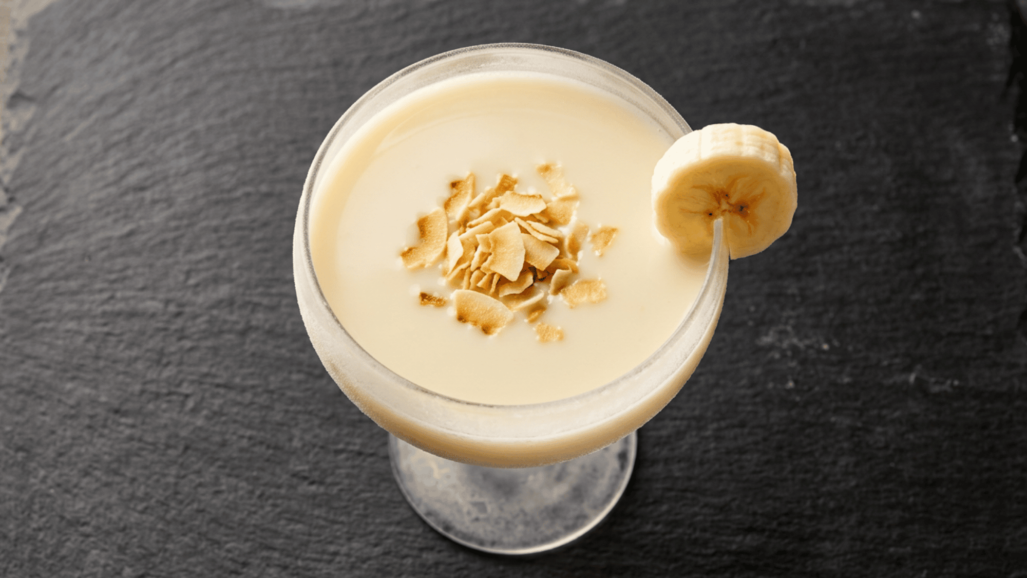 Creamy banana cocktail in a frosted coupe glass topped with toasted coconut flakes and a fresh banana slice