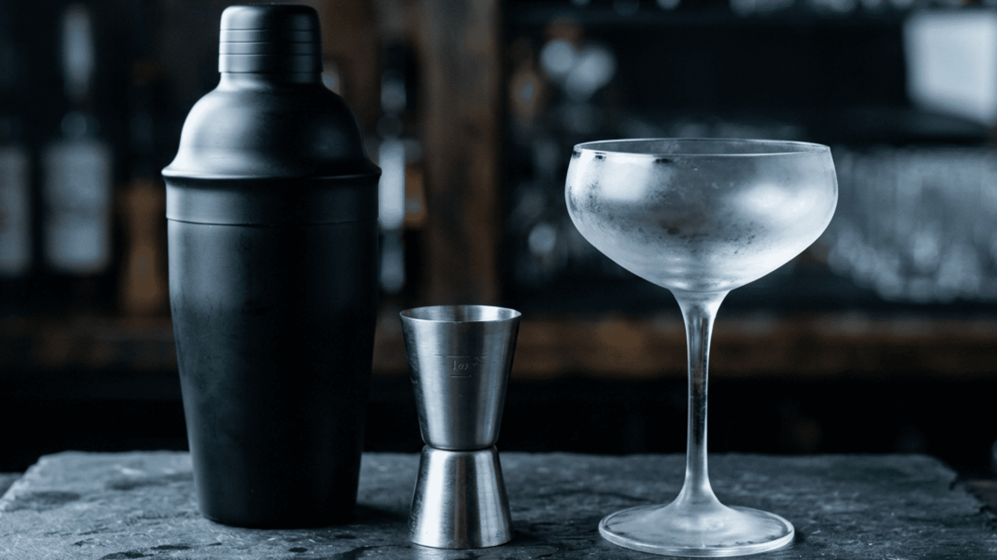 Black cocktail shaker, metal jigger, and chilled coupe glass neatly arranged on a bar counter before mixing