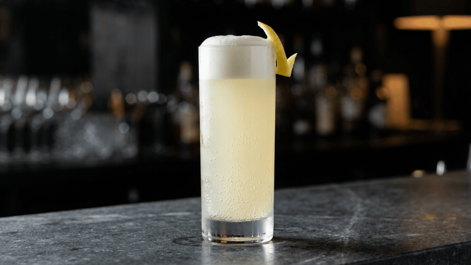Gin fizz with foam and lemon peel in a tall glass on a dark bar counter