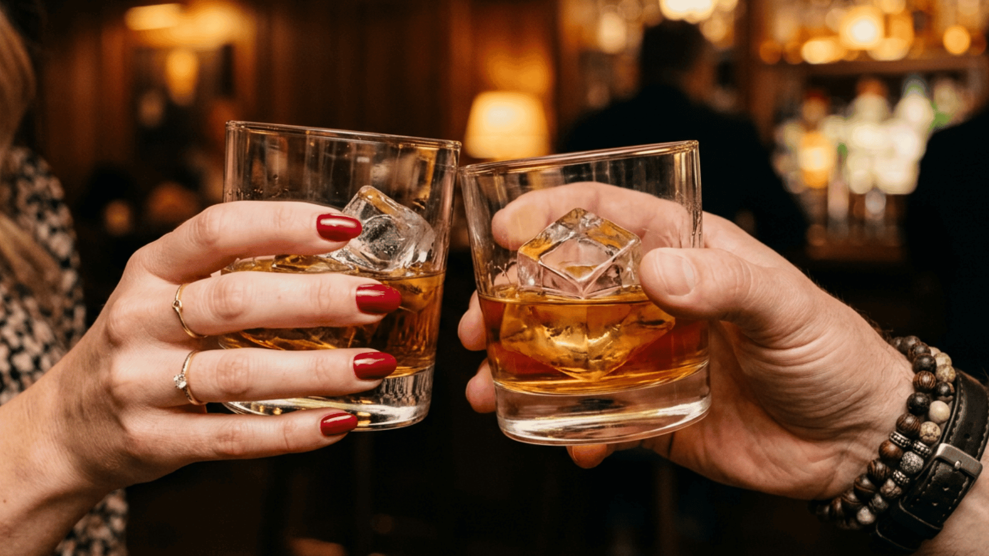Two people clinking glasses of whiskey on the rocks in a warm, cozy bar setting