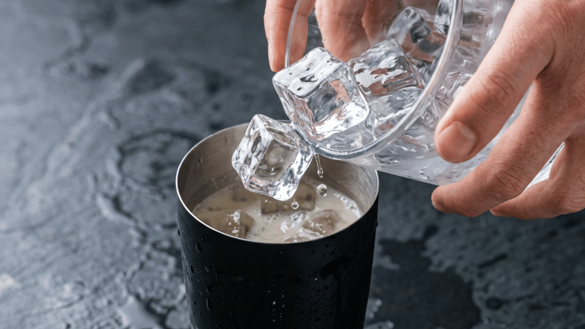Ice cubes being poured into a cocktail shaker filled with creamy banana cocktail ingredients on a stone countertop