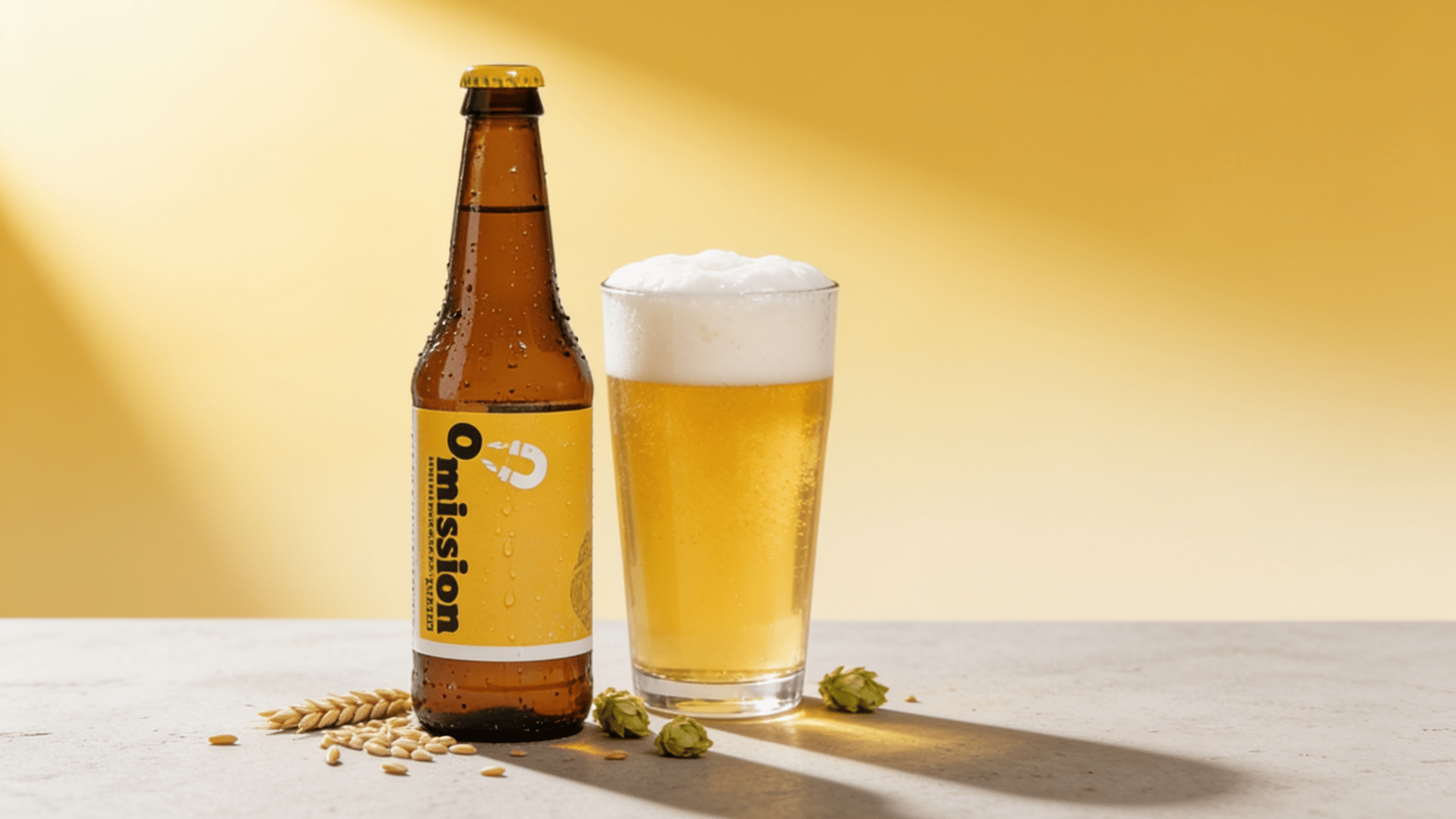 Omission Lager amber bottle beside a pint glass of crisp pale golden lager with barley grains on a concrete surface with a mustard yellow background.