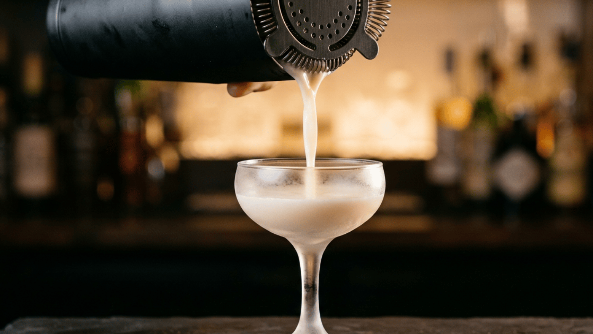 Creamy banana cocktail being strained from a shaker into a chilled coupe glass with a softly blurred bar background