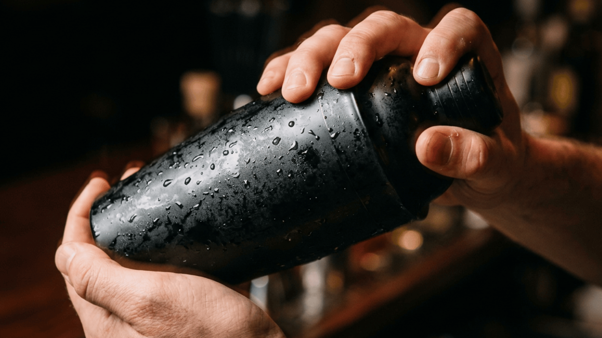 Hands vigorously shaking a chilled black cocktail shaker covered in condensation on a warm bar backdrop