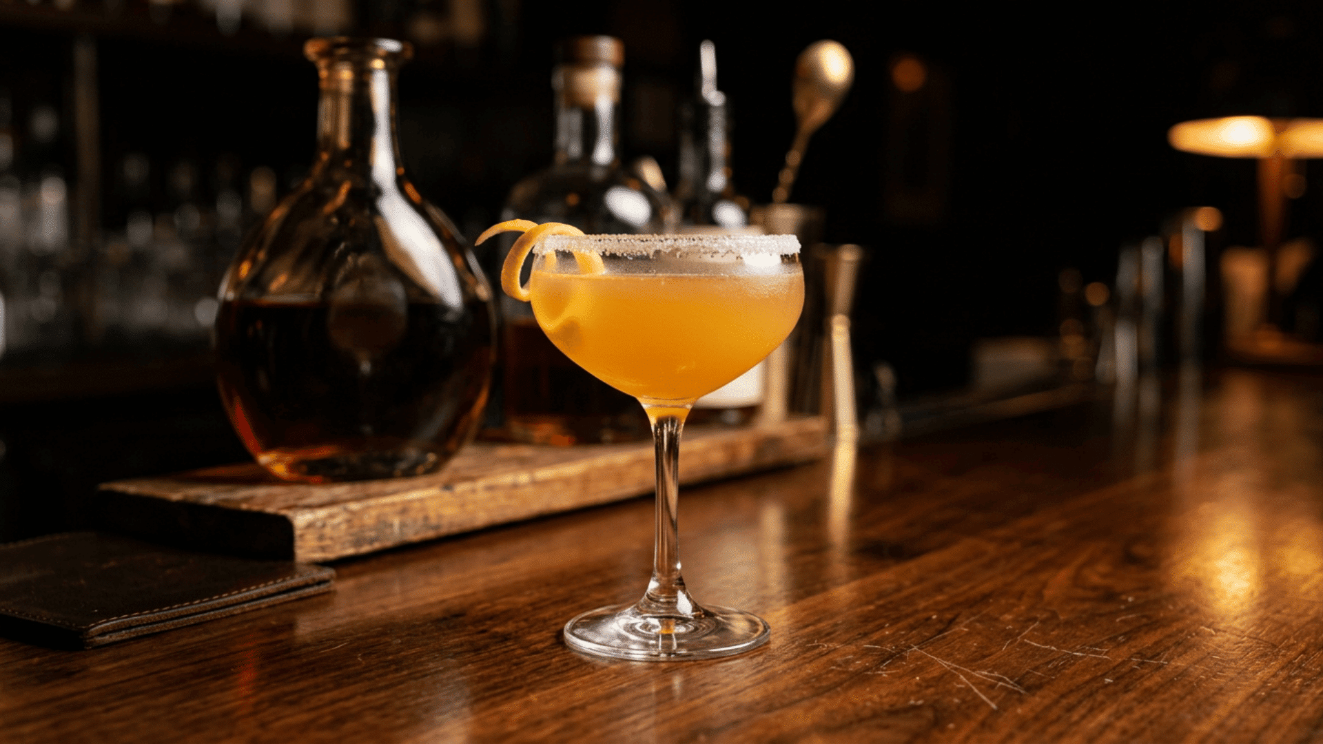 Sidecar cocktail with sugared rim and lemon twist on a wooden bar counter.