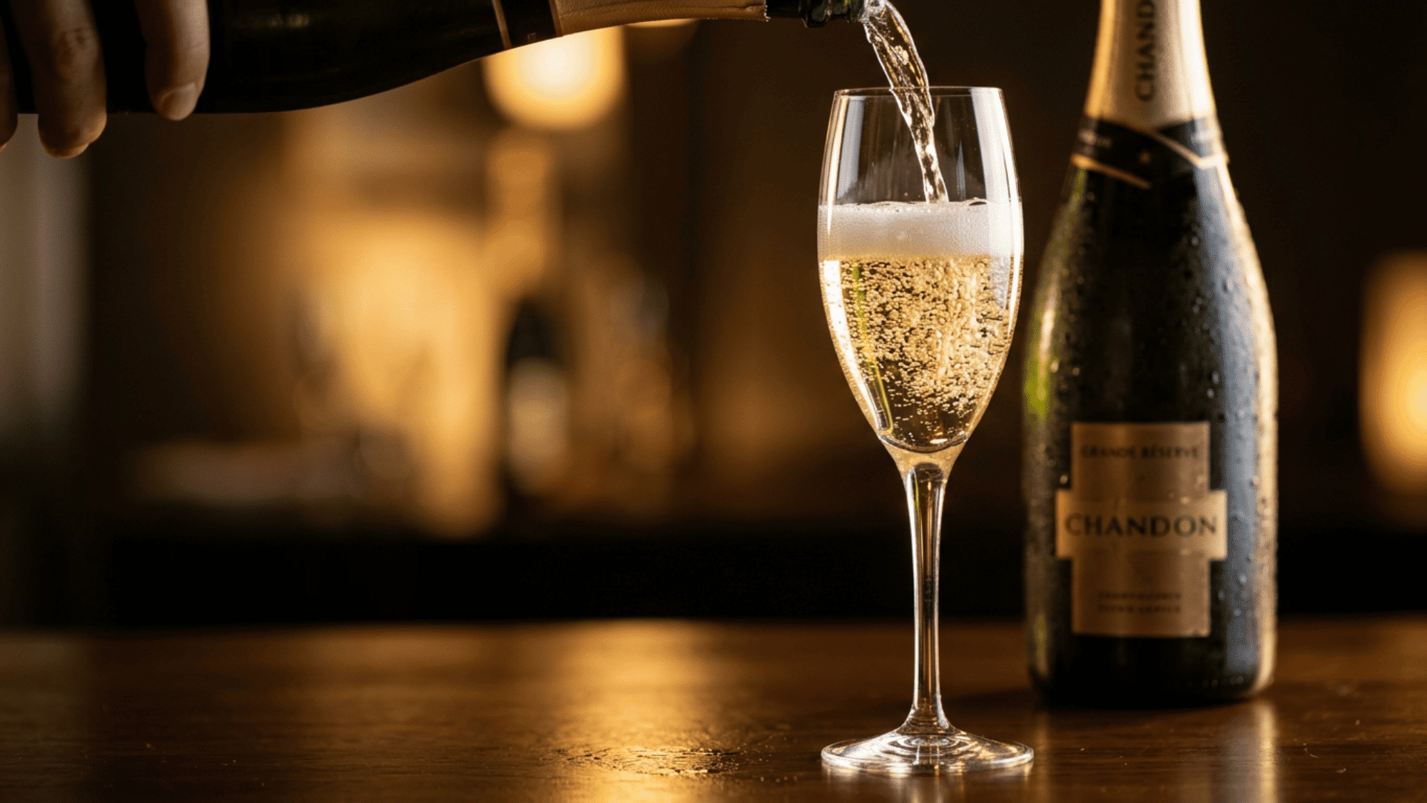 Sparkling wine being poured into a flute beside bottle in warm golden bar lights