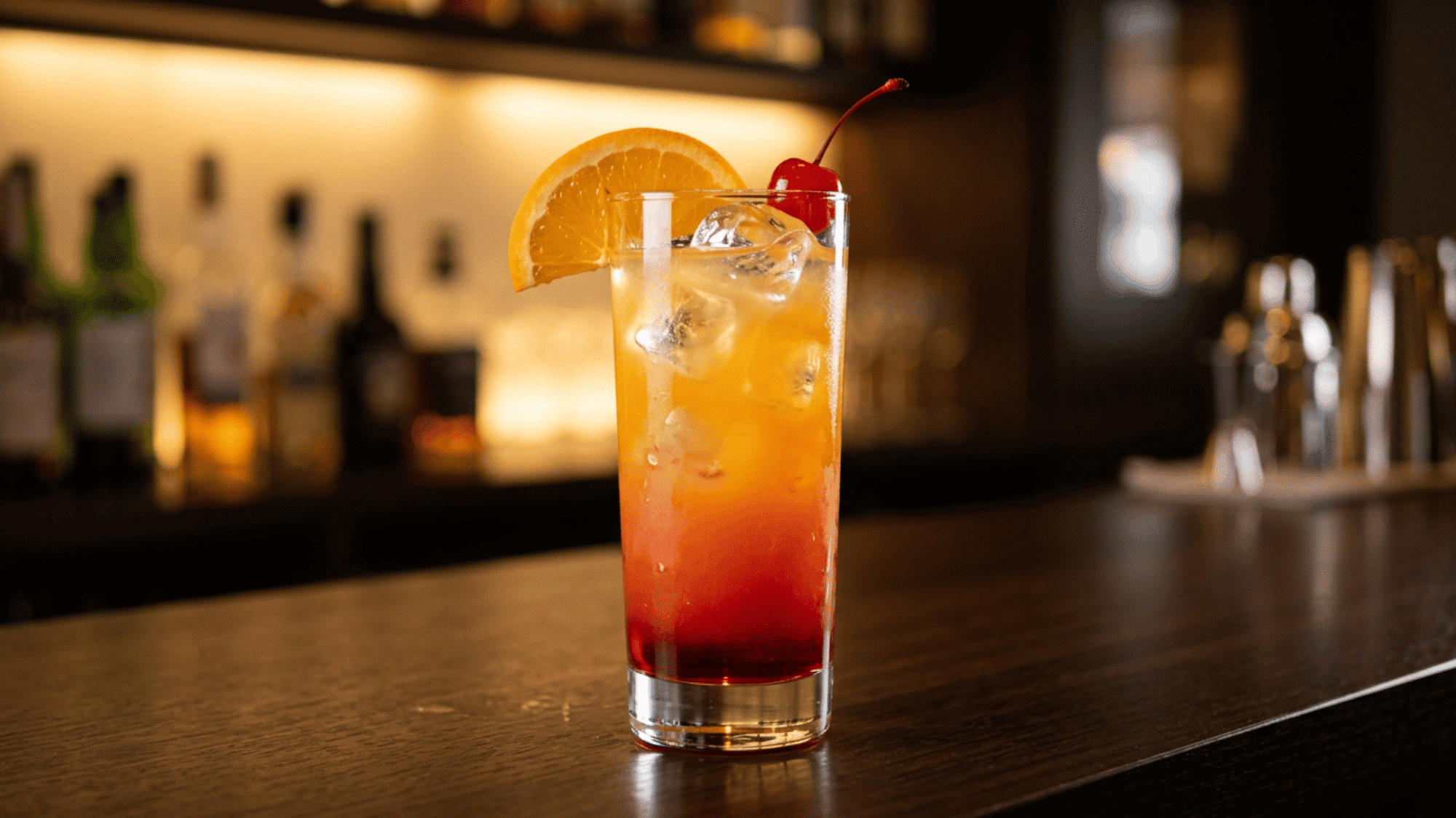 Tequila Sunrise cocktail with orange, cherry, ice, and red-orange layers on a bar.