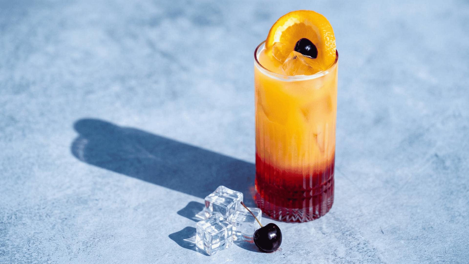 Tequila Sunrise cocktail with red and orange layers, ice, orange slice, and cherry.