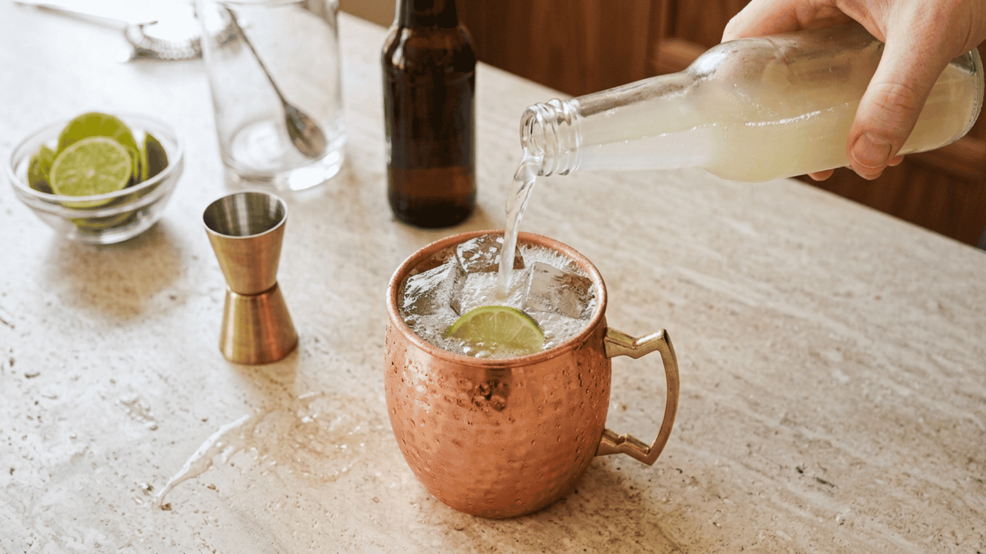 Hand pouring ginger beer into an iced Moscow Mule in a copper mug.