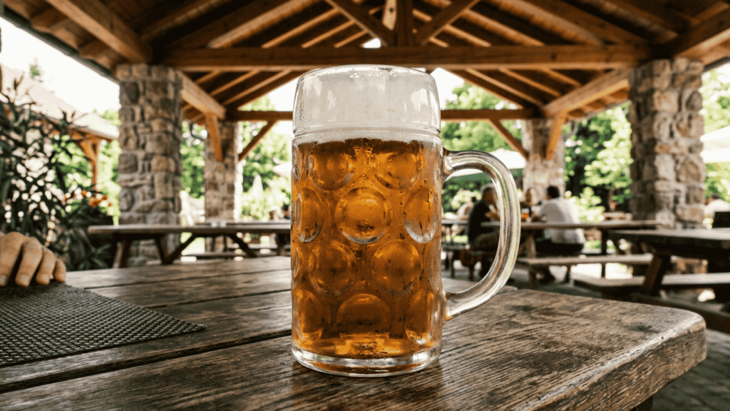 Golden farmhouse ale in a frothy beer mug served on a rustic wooden table inside a cozy beer garden.