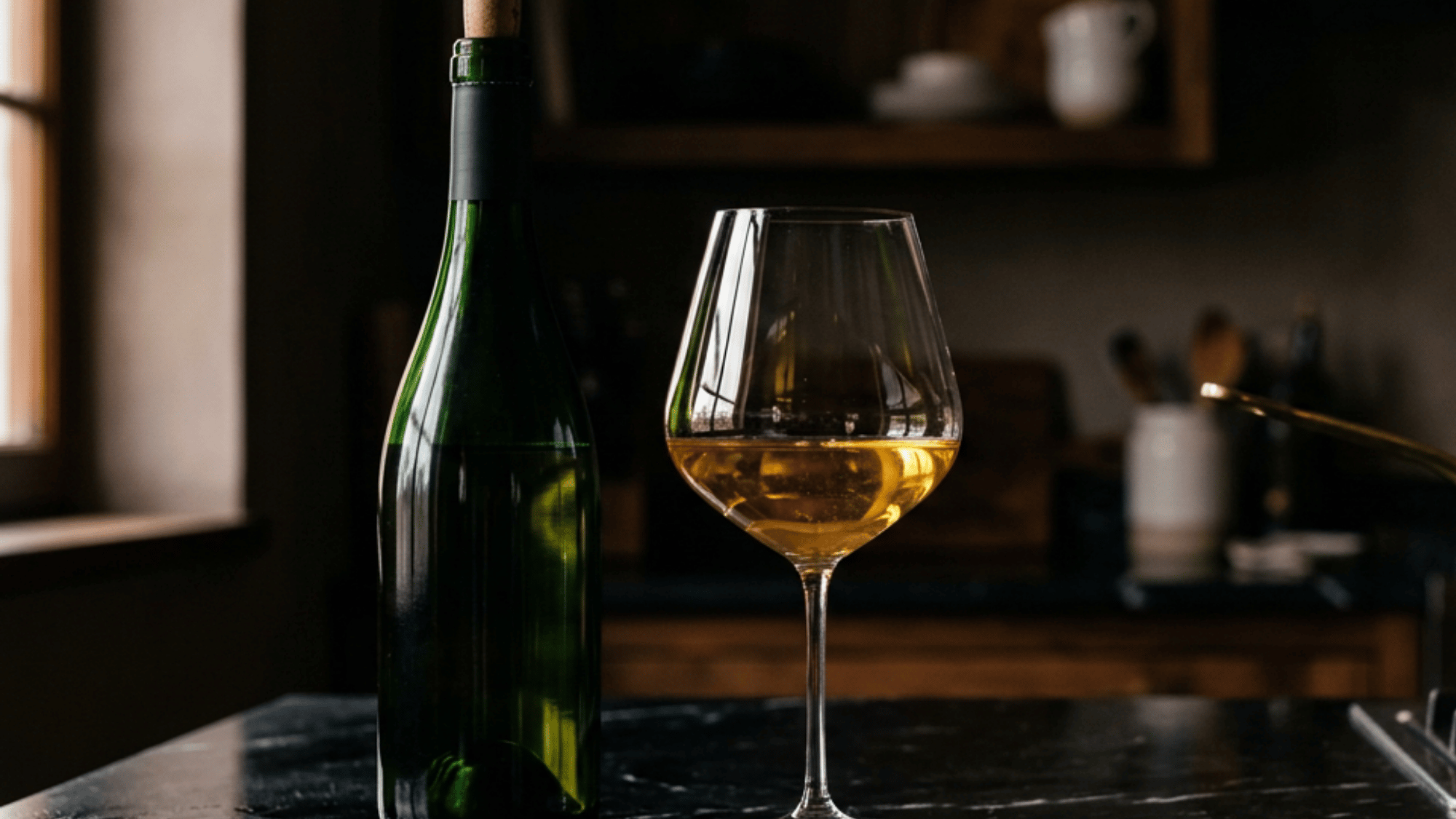 Green wine bottle and glass of White Burgundy on a dark kitchen countertop.
