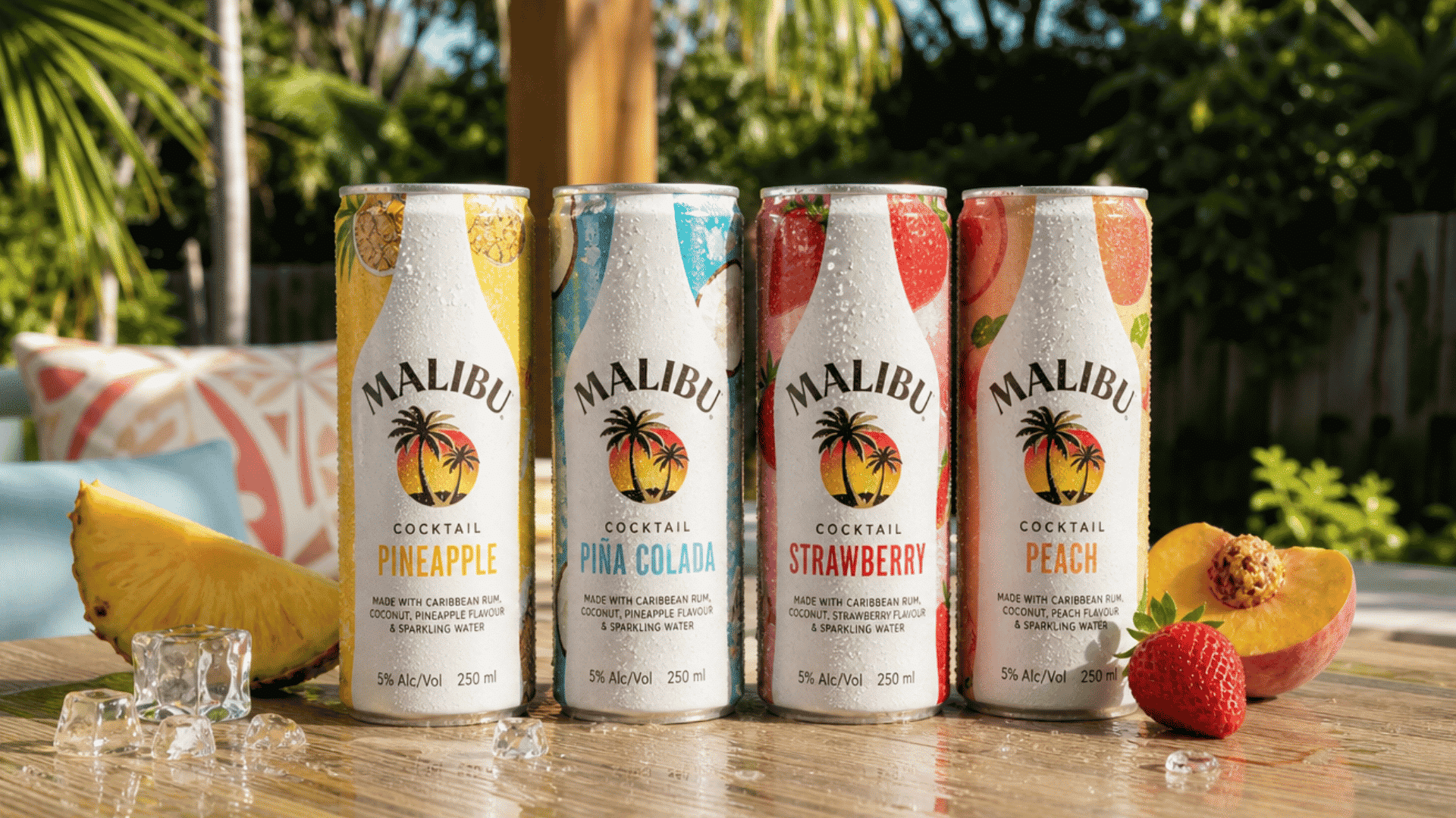 four chilled Malibu cocktail cans in pineapple, piña colada, strawberry, and peach flavors on a tropical patio table.