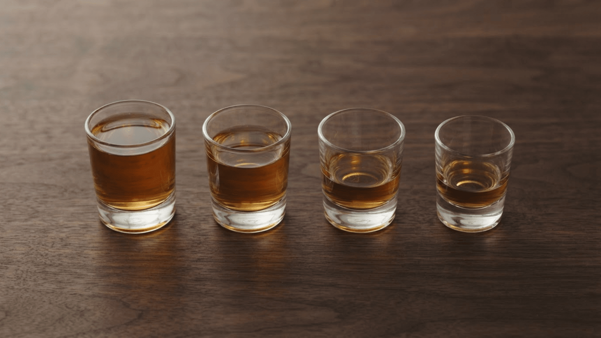 four shot glasses with different amounts of amber liquor arranged on a wooden surface