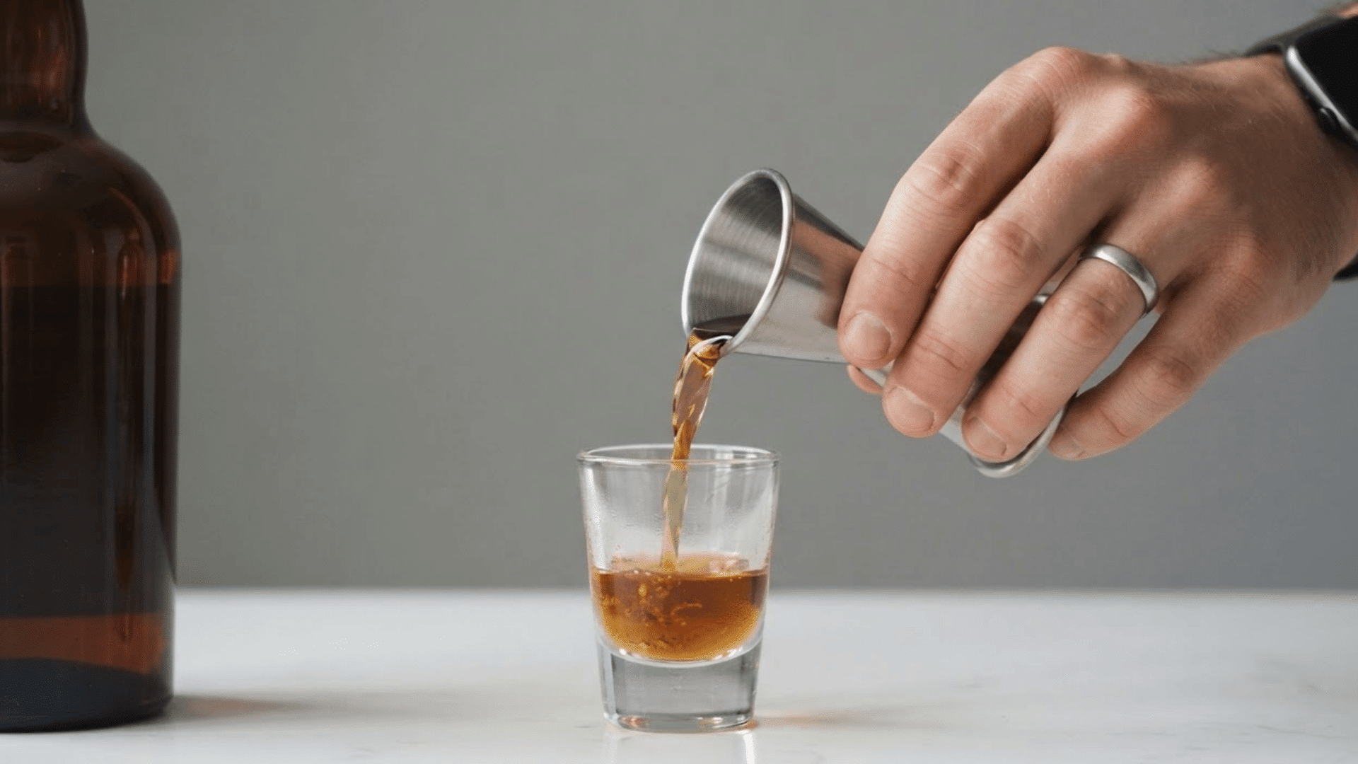hand pouring liquor from a metal jigger into a glass with ice and fresh mint