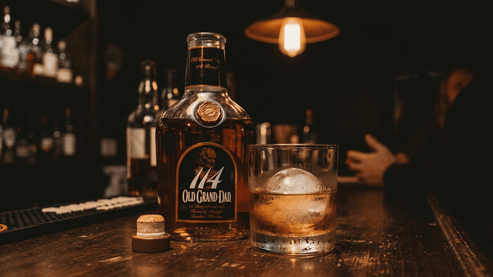 old grand dad 114 bourbon bottle beside a glass with a round ice ball in dim bar lighting at night