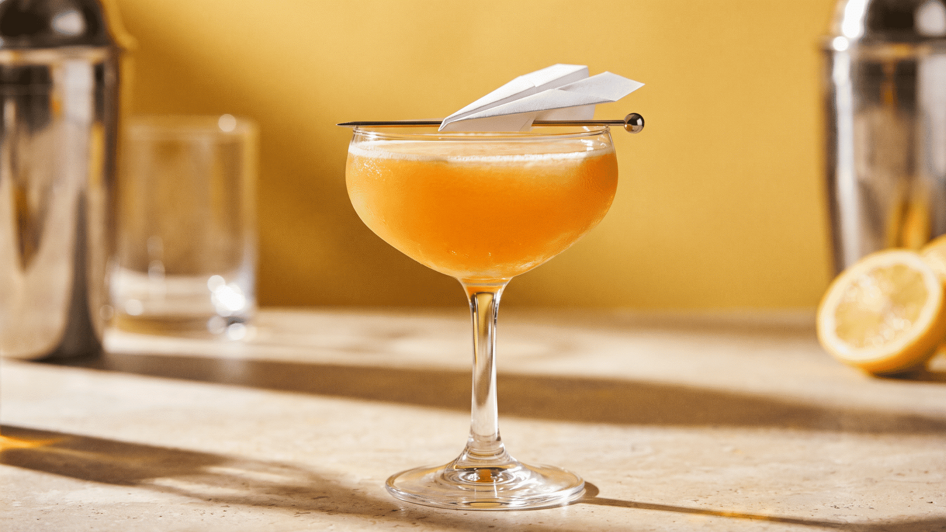 Orange cocktail in coupe glass with paper plane garnish on a sunlit bar.