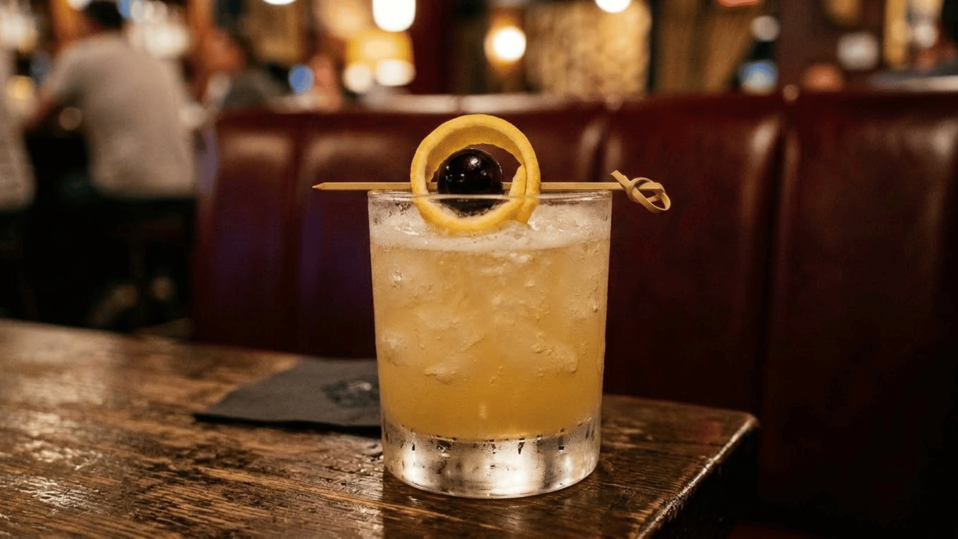 saturn cocktail with pale golden foam, garnished with a citrus peel ring and cherry skewered on a toothpic