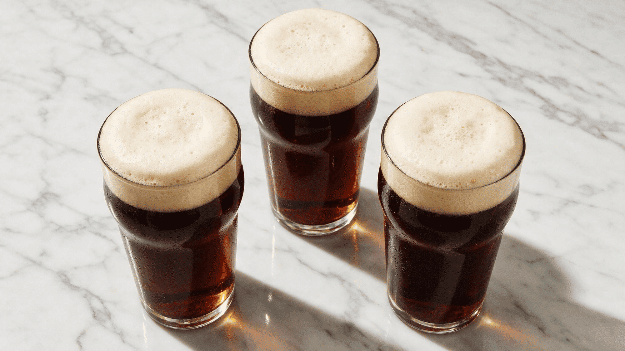 Three different sized glasses of dark stout beer arranged side by side on a white marble surface