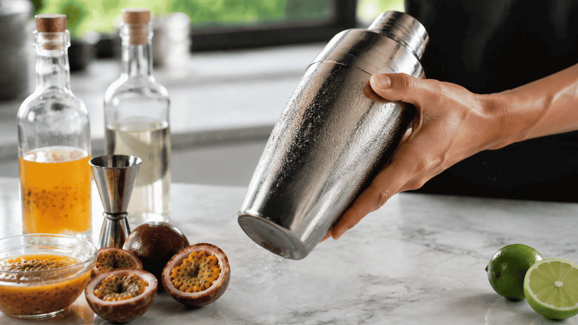 Bartender shaking a chilled stainless steel cocktail shaker with fresh ingredients on a marble countertop.