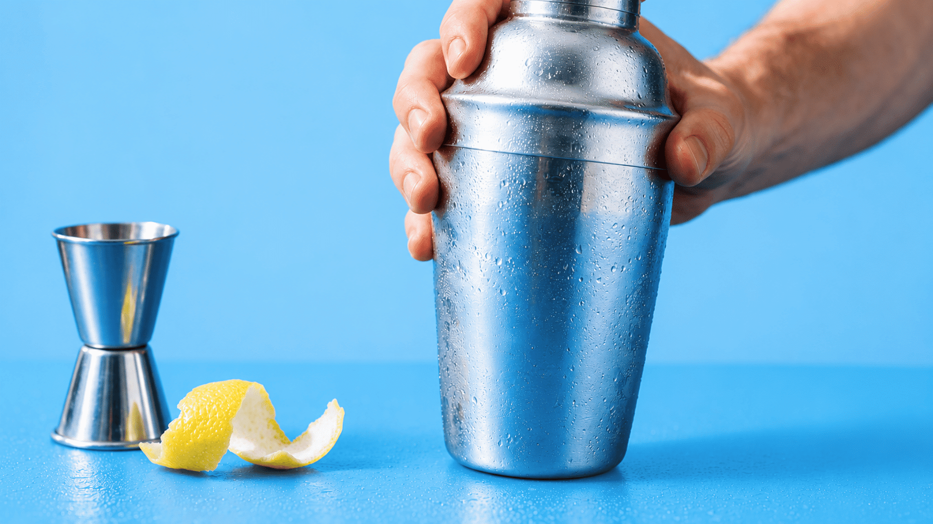 Hands shaking a chilled stainless steel cocktail shaker beside a metal jigger and fresh lemon peel on a dark bar counter.
