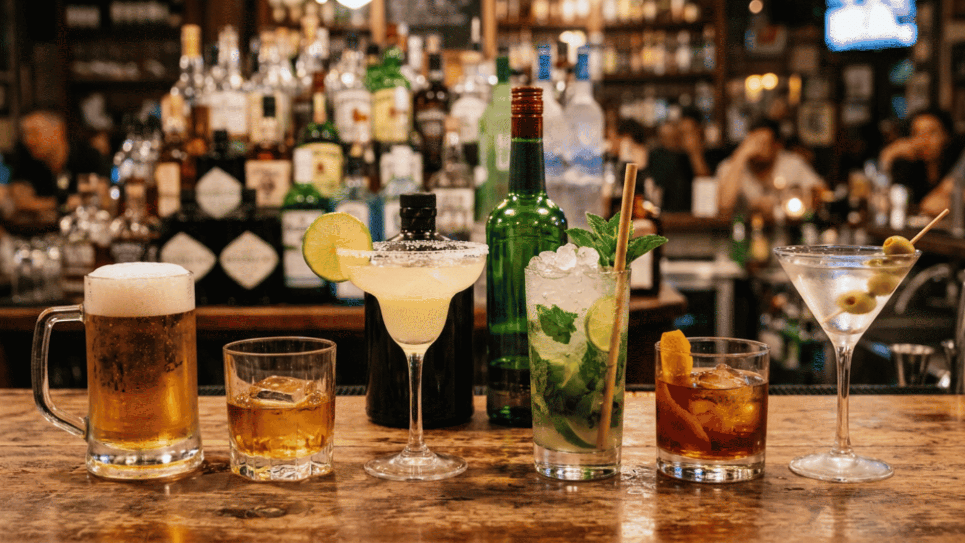 beer, whiskey, margarita, mojito, old fashioned, and martini lined up on a wooden bar with liquor bottles behind them