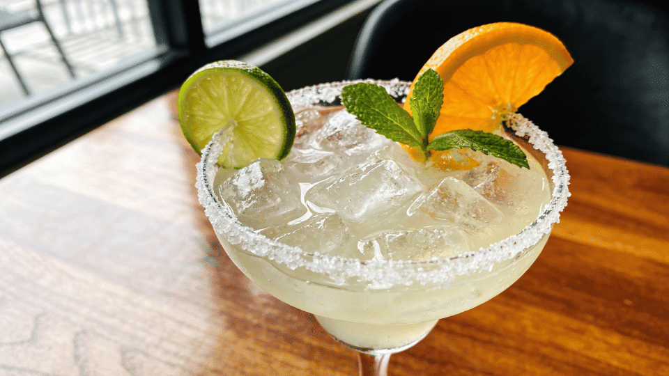 virgin margarita in a salted-rim glass with lime, orange, mint and ice served on a wooden table by a window