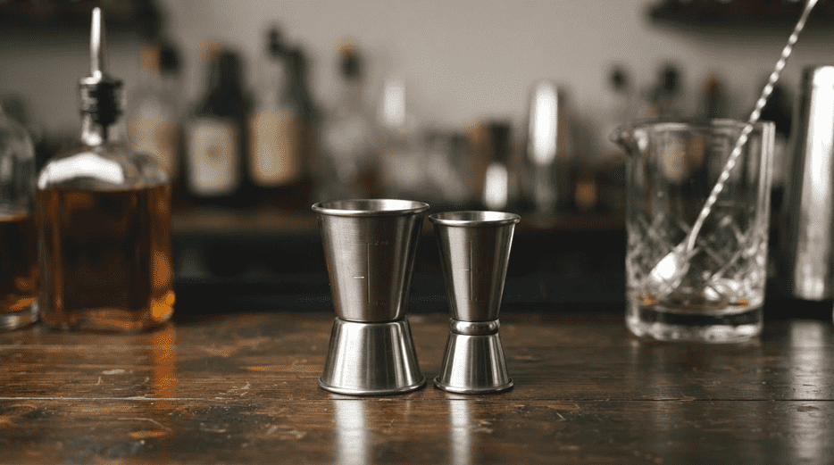 Stainless steel double jigger beside a mixing glass and liquor bottle on a wooden bar